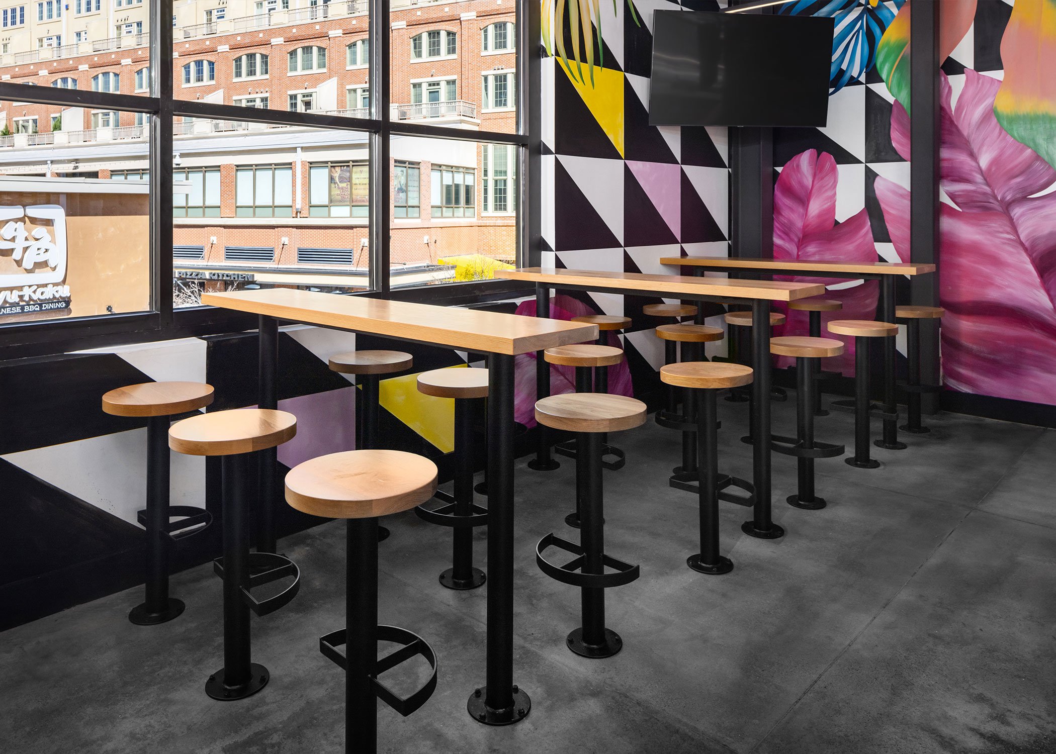 Three bar height tables with 6 sally barstools with footrings lining a corner with large pink leaves painted in a mural on the wall.