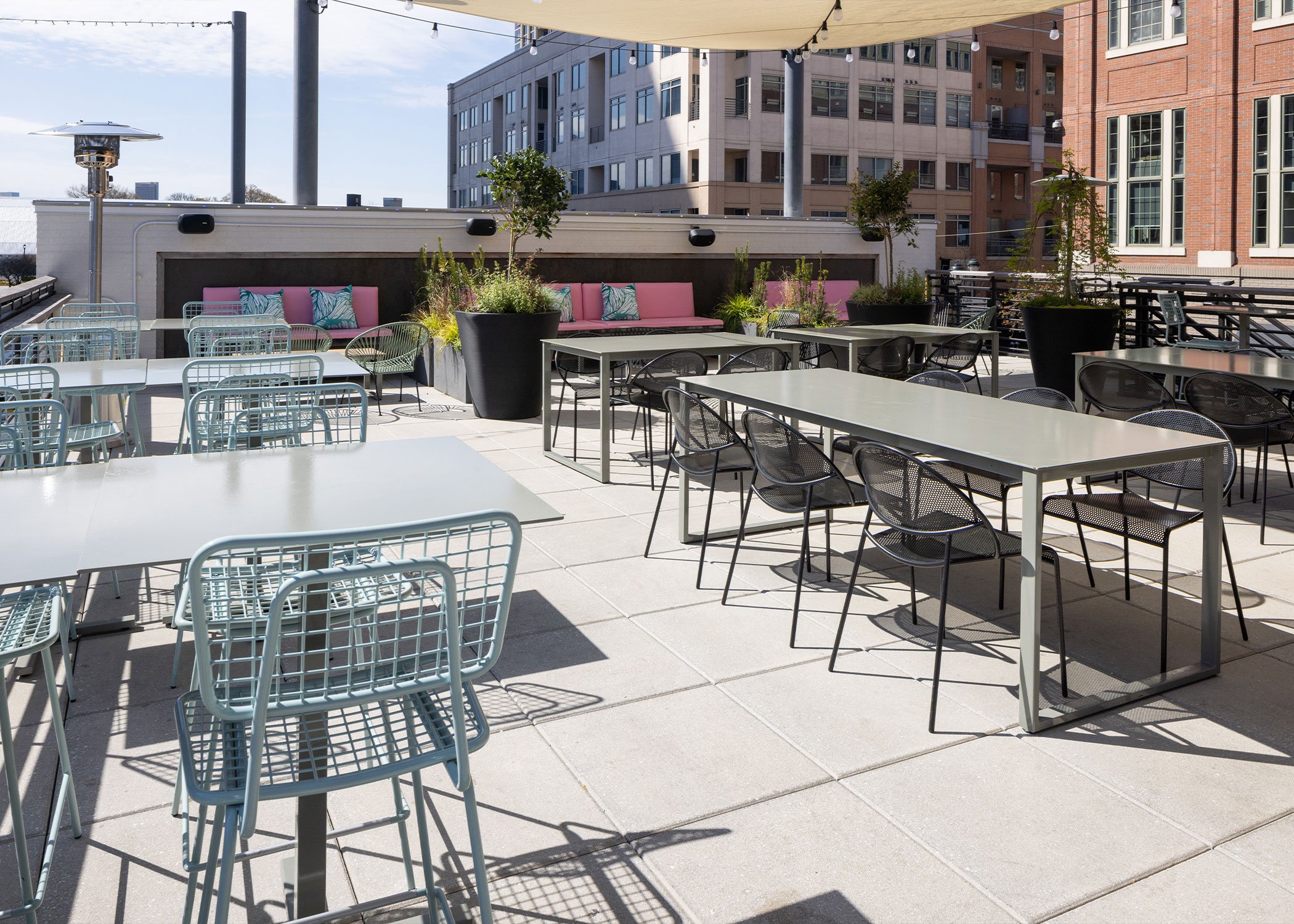 A patio at Azotea with many dusty blue opla chairs and black Hula chairs at steel tables under wide canopies and string lights. Pink couches against the wall in the background.