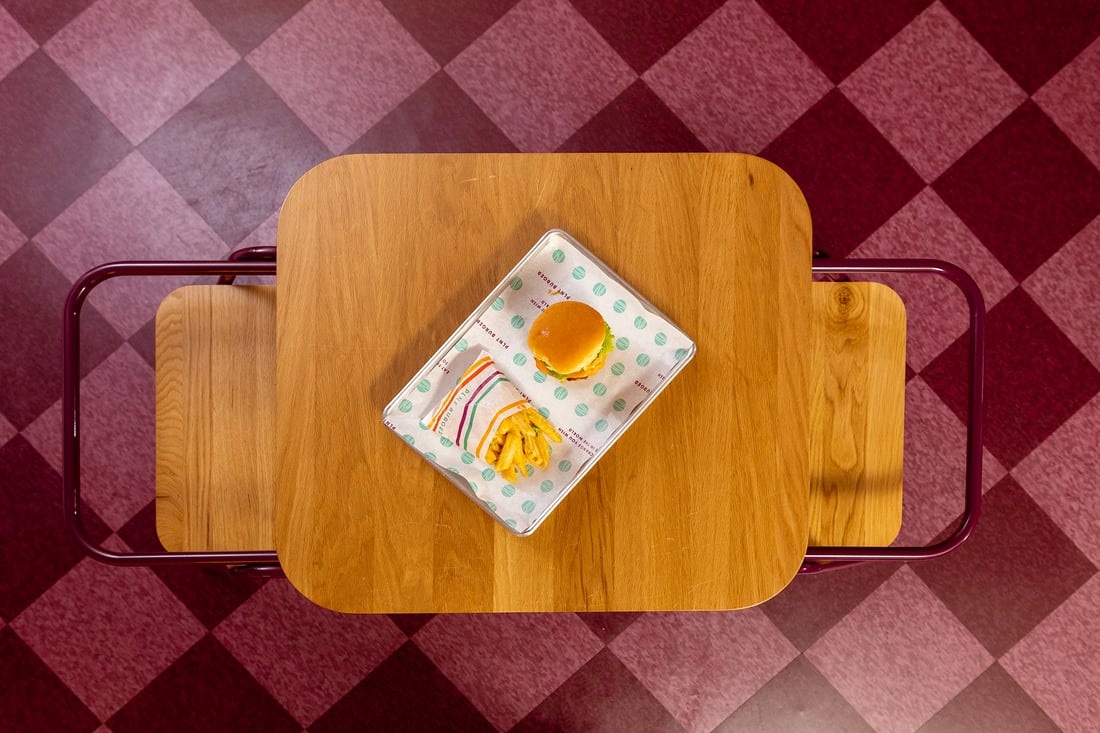 Overhead view of wood top with a burger and fries in the center surrounded by magenta checkered floors.
