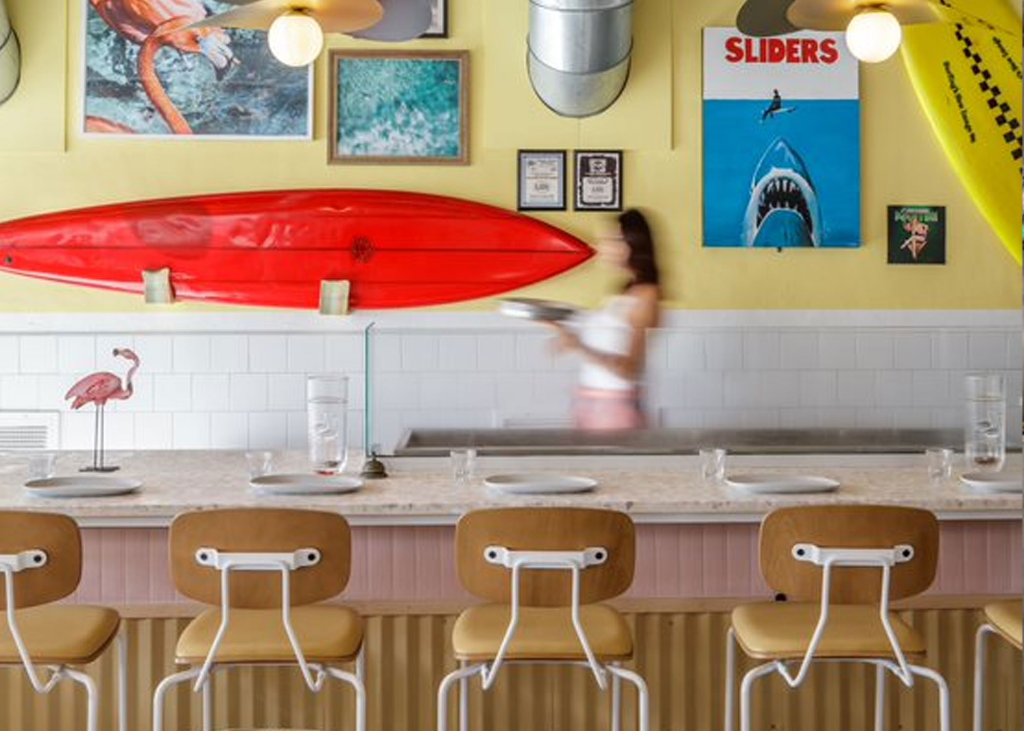 Reece barstools with tan upholstered seats, wood backs, and white steel frames line a bar set with tables. A server is passing behind it, blurred by motion. The wall is yellow with a surfboard, a fake Jaws poster with Slider's in red text and other beachy art pieces.