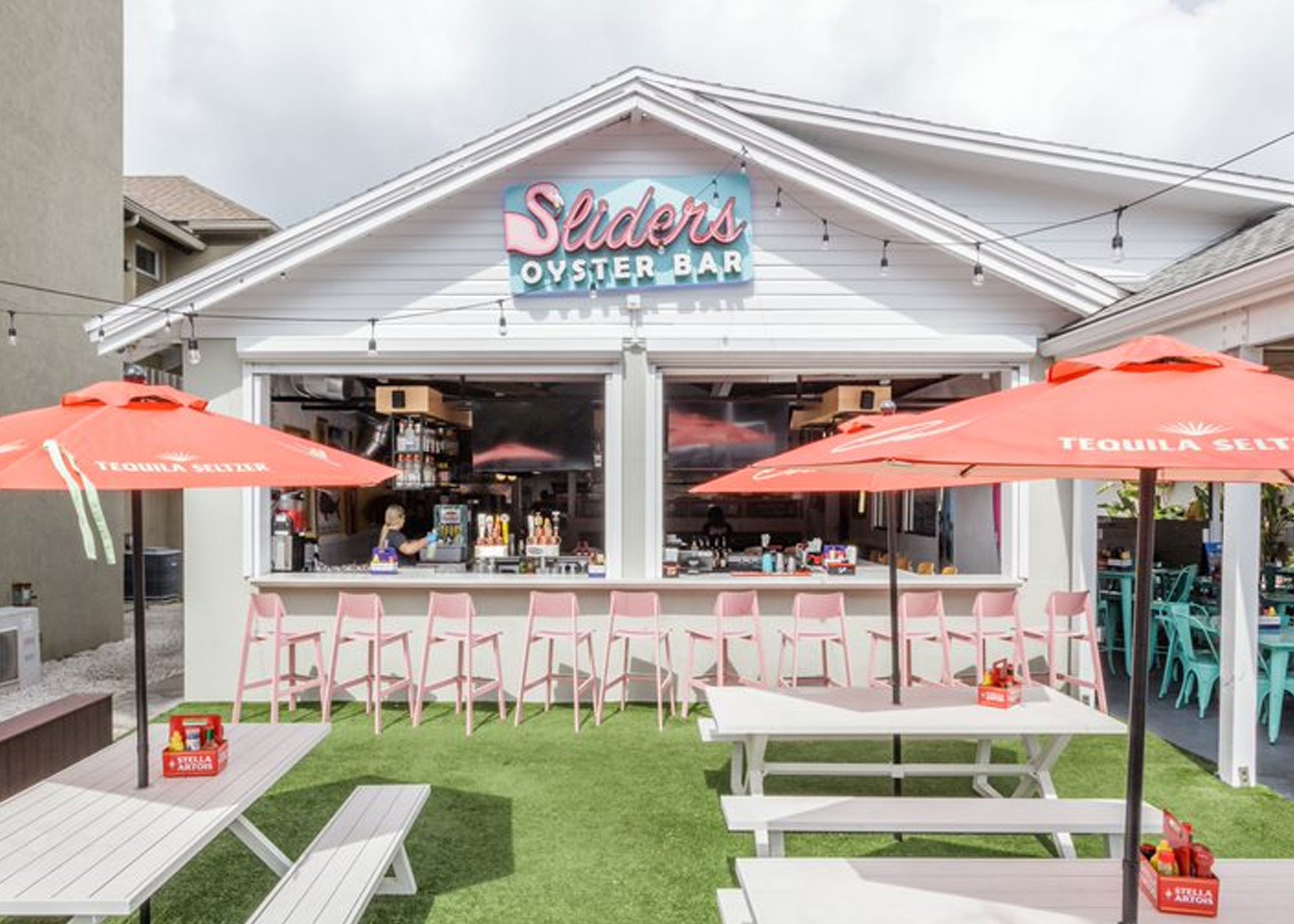 Slider's exterior has a bar in front with pink Sadie II outdoor chairs lined at it. Picnic tables sit on green turf with red umbrellas. A sign with the restaurants name hangs over the bar under the point of the roof.