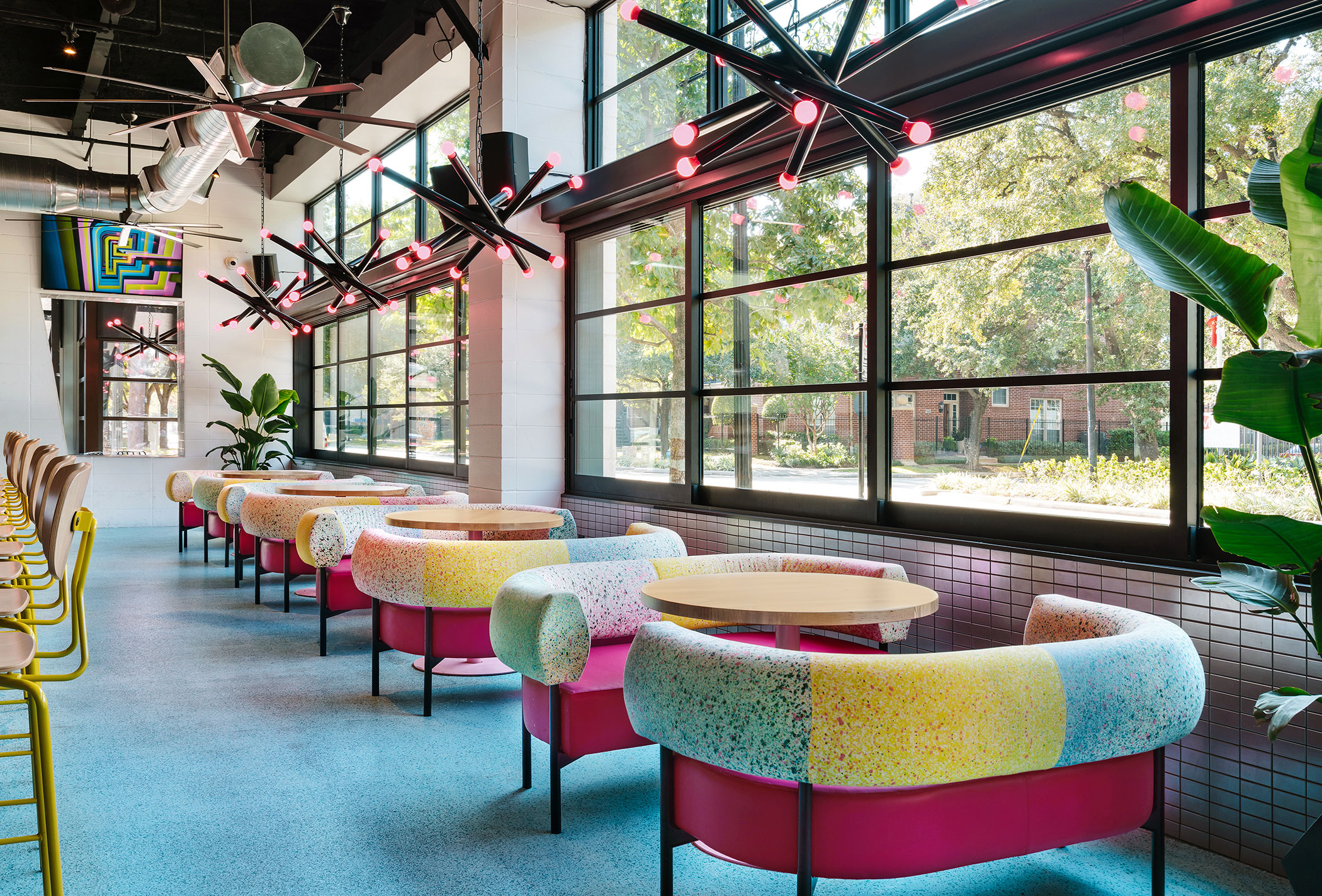 Sets of colorful sofa seating around a hand full of tables lining windows at a restaurant, modern chandelier lighting overhead.