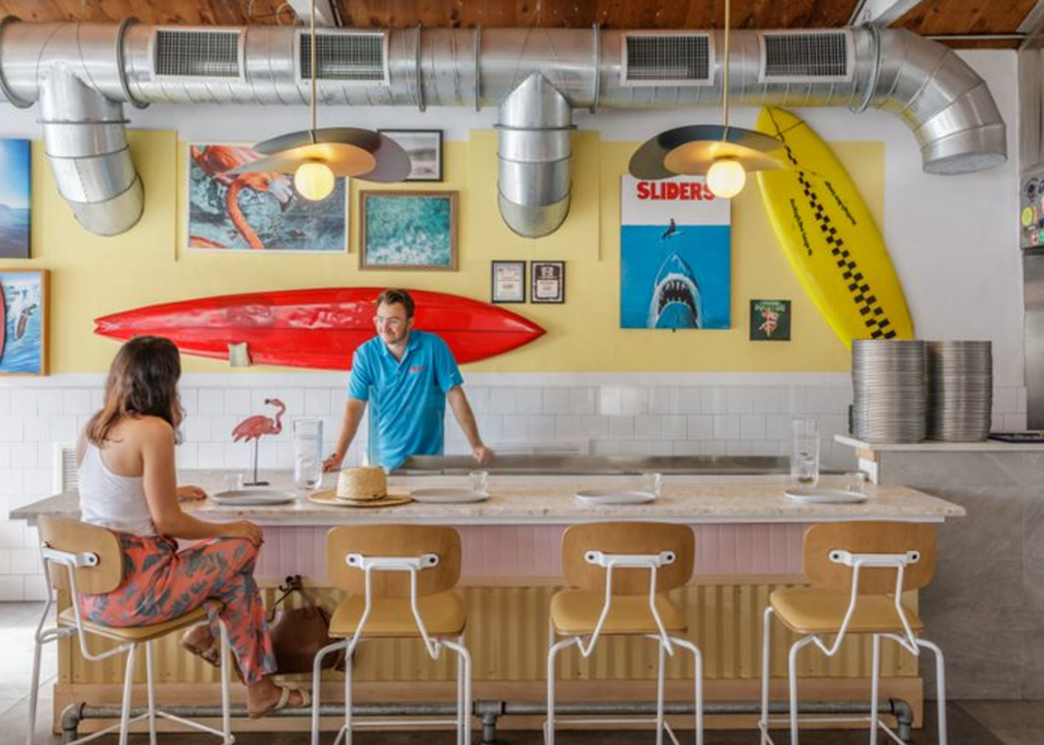 A woman sits at the bar in a Reece barstool, facing a bartender who leans against the bar. The wall behind them is yellow with beachy wall art and surfboards hanging on it. 