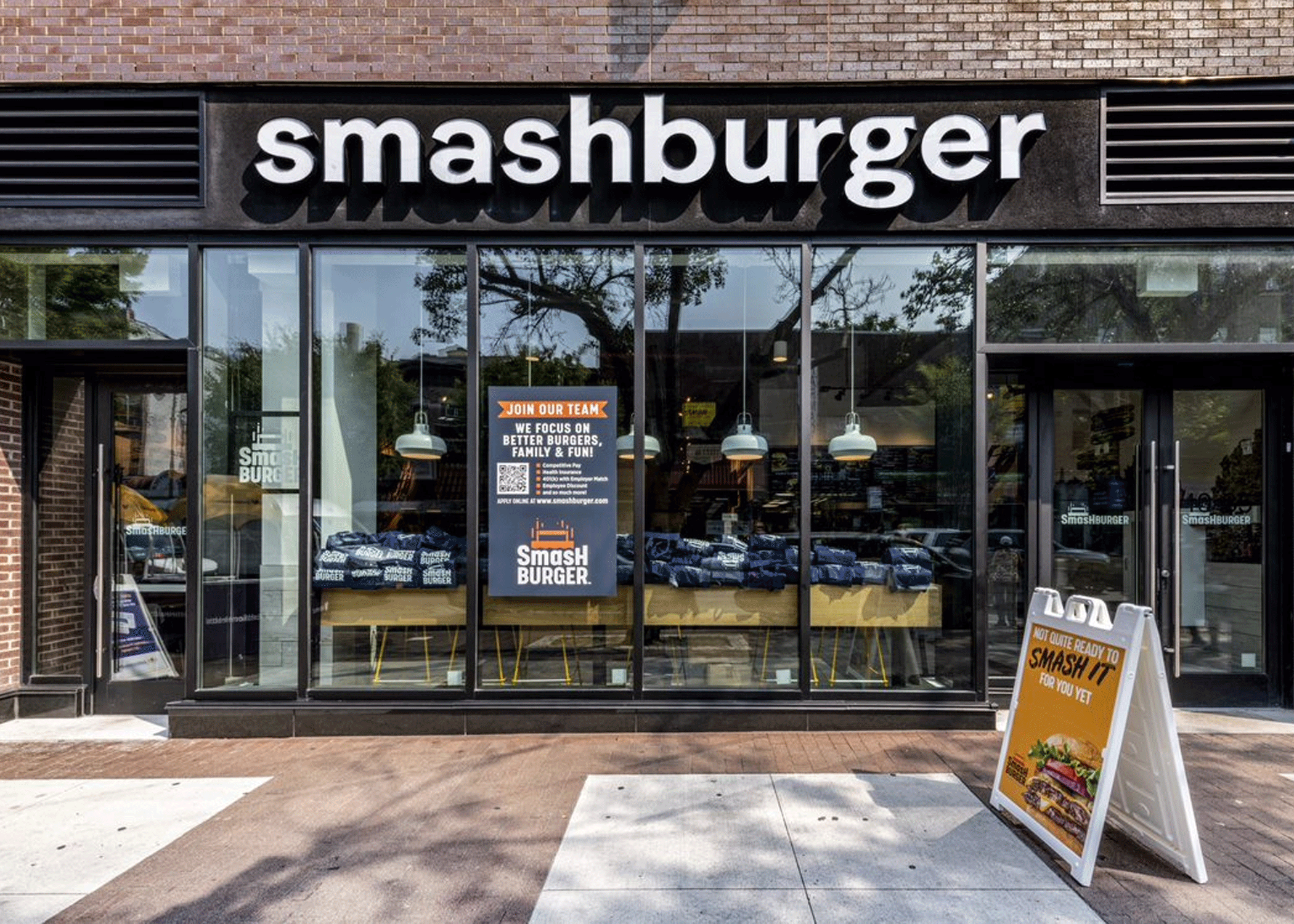 Storefront with glass windows on a wide sidewalk with a sign posted outside that features a burger. Big text above the window reads Smashburger.