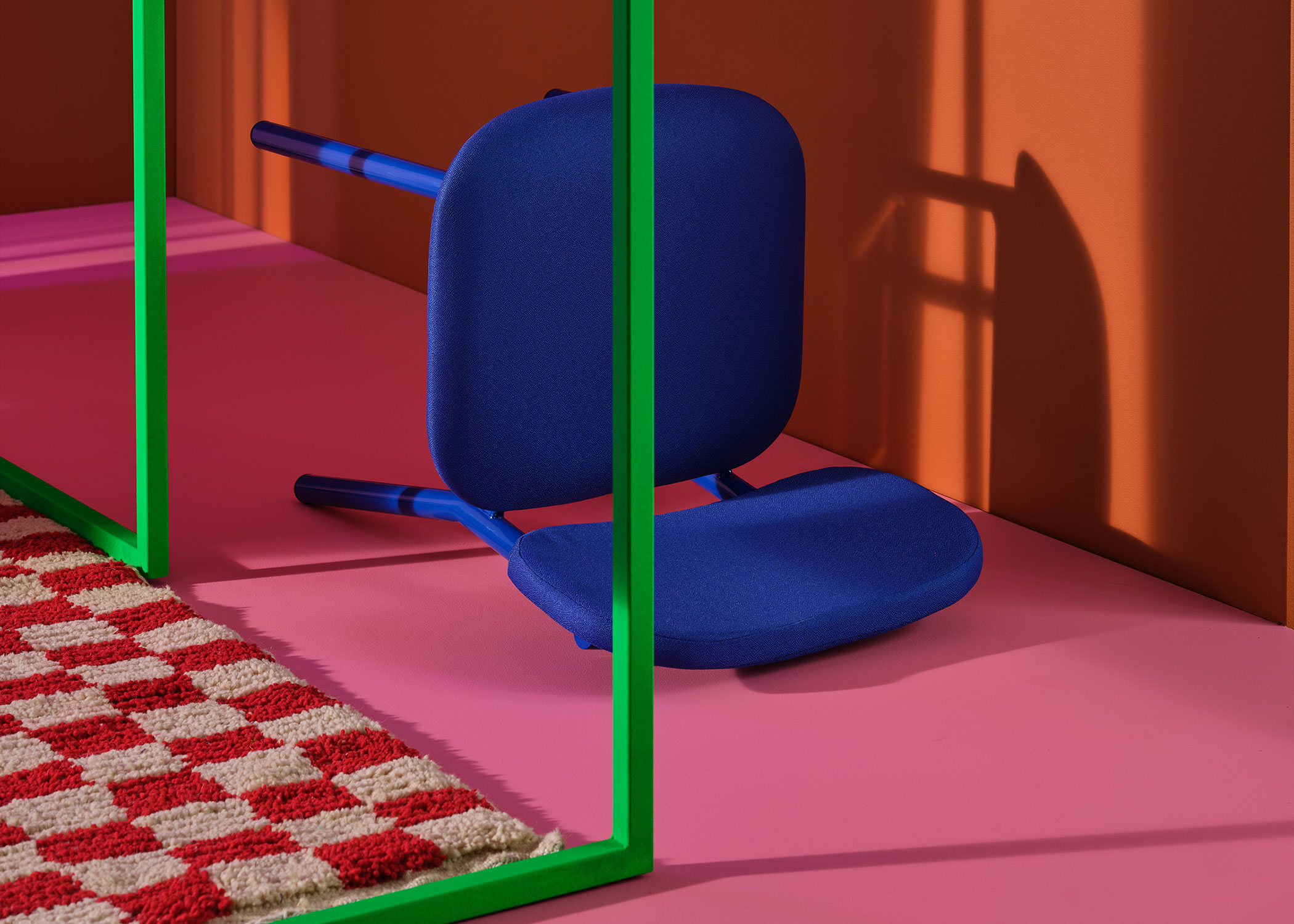 Bright Blue Ferdinand Chair laying on the floor on the back. Floor is bright pink, wall is orange. A green steel frame cuts across the chair. A white and red checkered rug sits under the frame.