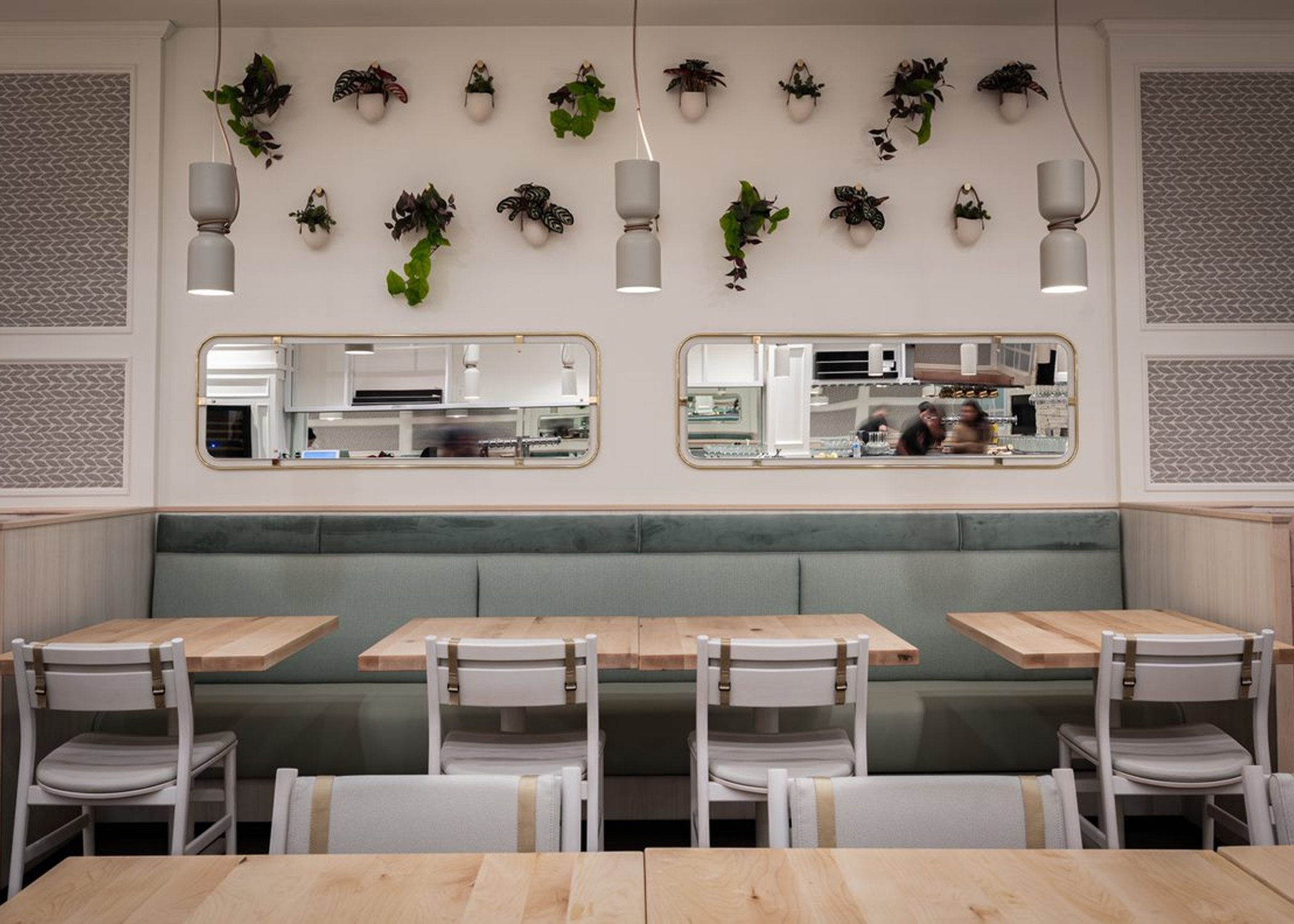 Wall with two rows of mounted plants above two windows into the kitchen. Pale blue booths below the mirrors with tables and Sigsbee chairs in a pale finish with khaki buckles, fully upholstered.
