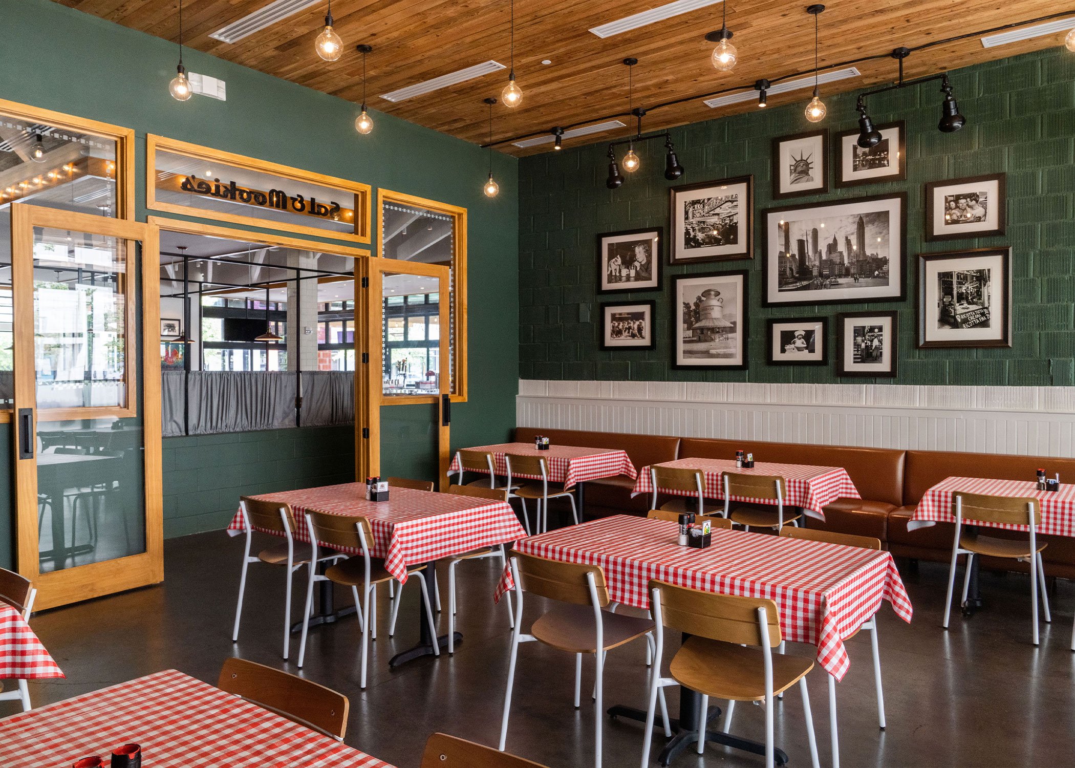 Restaurant interior with Sherman chairs at tables covered with classic red plaid table cloths. Green walls with warm natural wood details, framed black and white images on a gallery wall